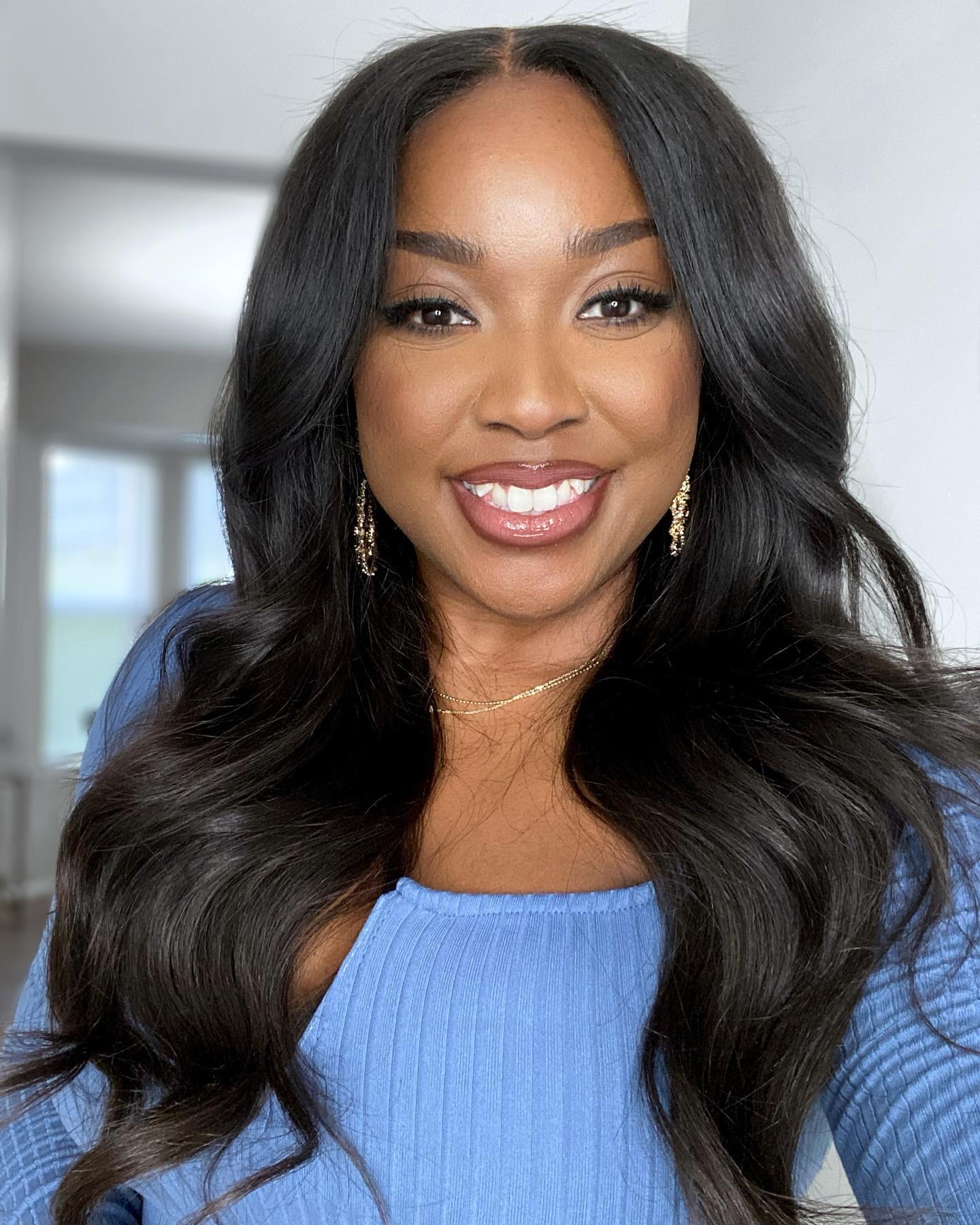 Jaslyn smiling in a blue top with softly waved hair and gold earrings